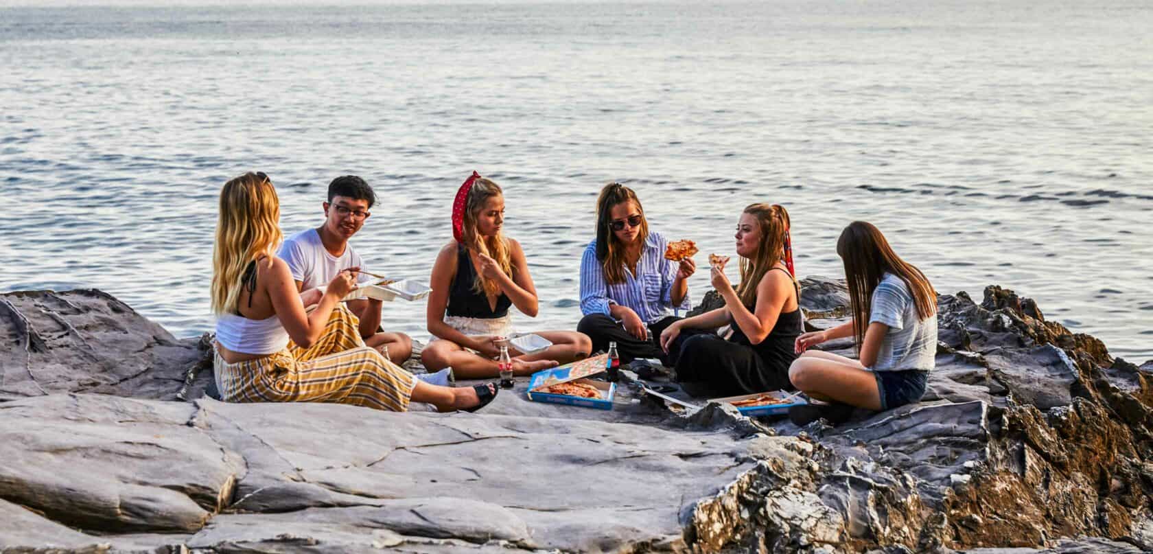 Friends sitting on a rock near the ocean eating food.